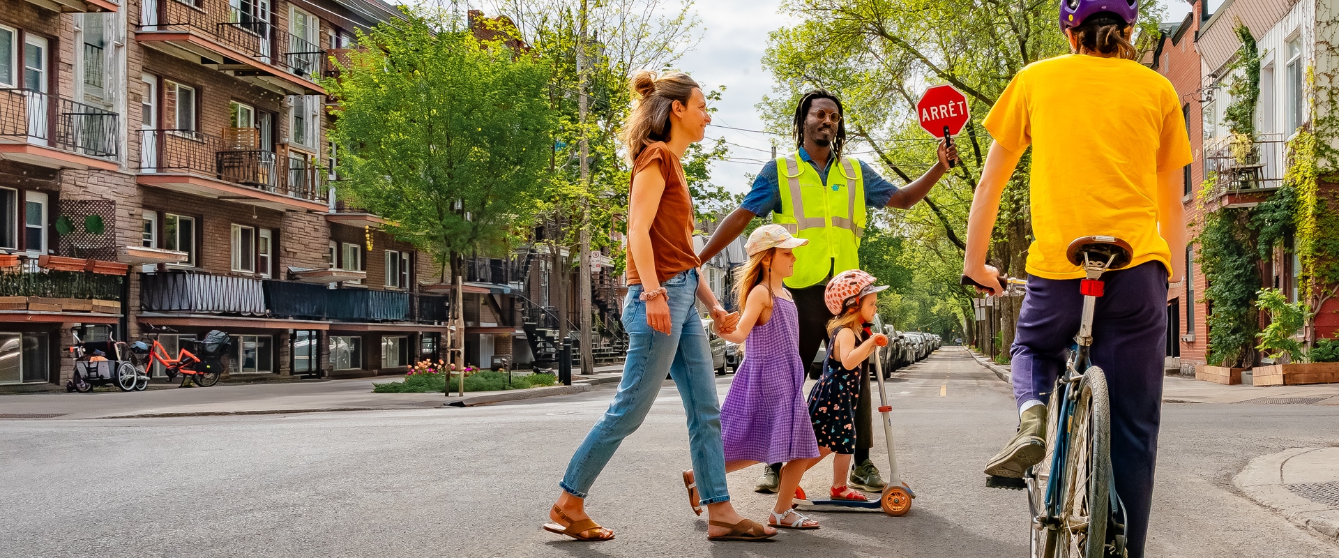 Scène à une intersection de rues bordées d'arbres, où un cycliste est arrêté avant l'intersection, afin de laisser traverser une mère et ses deux petites filles, dont une est en trottinette. Derrière elles, un brigadier scolaire sécurise le passage avec son panneau d'arrêt à la main. Un texte en surimpression précise « Piéton en vue, priorité absolue. »