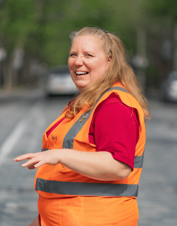 Portrait souriant de Danny Martel, une femme joviale aux cheveux blonds qui porte une veste de haute visibilité orange avec des bandes réfléchissantes.